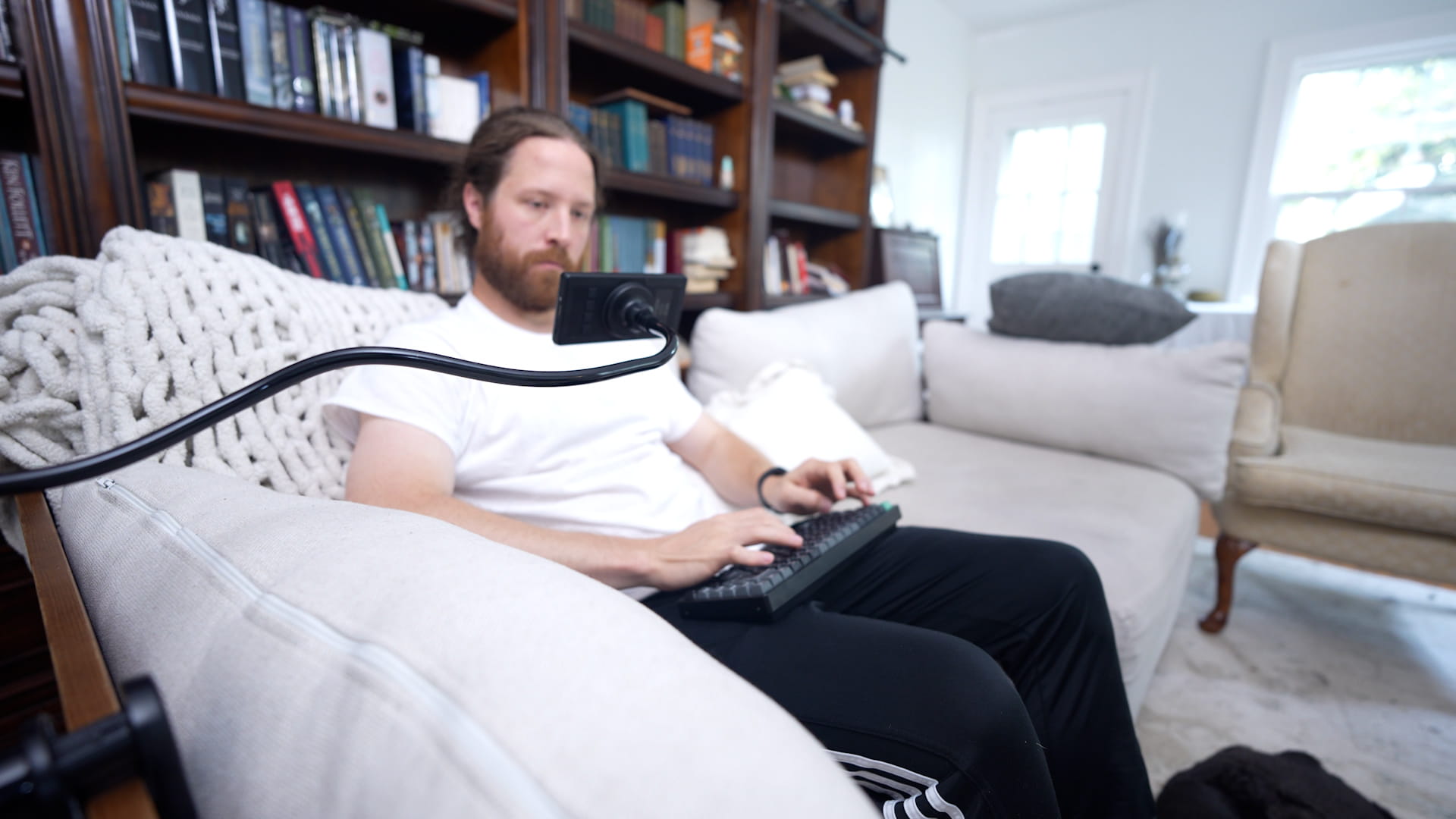 Writer on a couch typing on a Keychron K2 Ultra with the BYOK held in front of him by a gooseneck arm clamped to the sofa