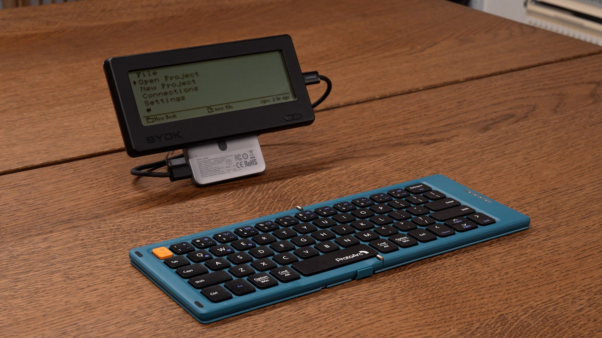 BYOK mounted on a charging stand with a USB-C cable, beside an unfolded travel keyboard